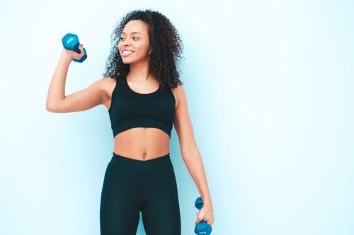 Fitness smiling black woman in sports clothing with afro curls hairstyle.She wearing sportswear. Young beautiful model with perfect tanned body.Female holding dumbbells in studio near light blue wall